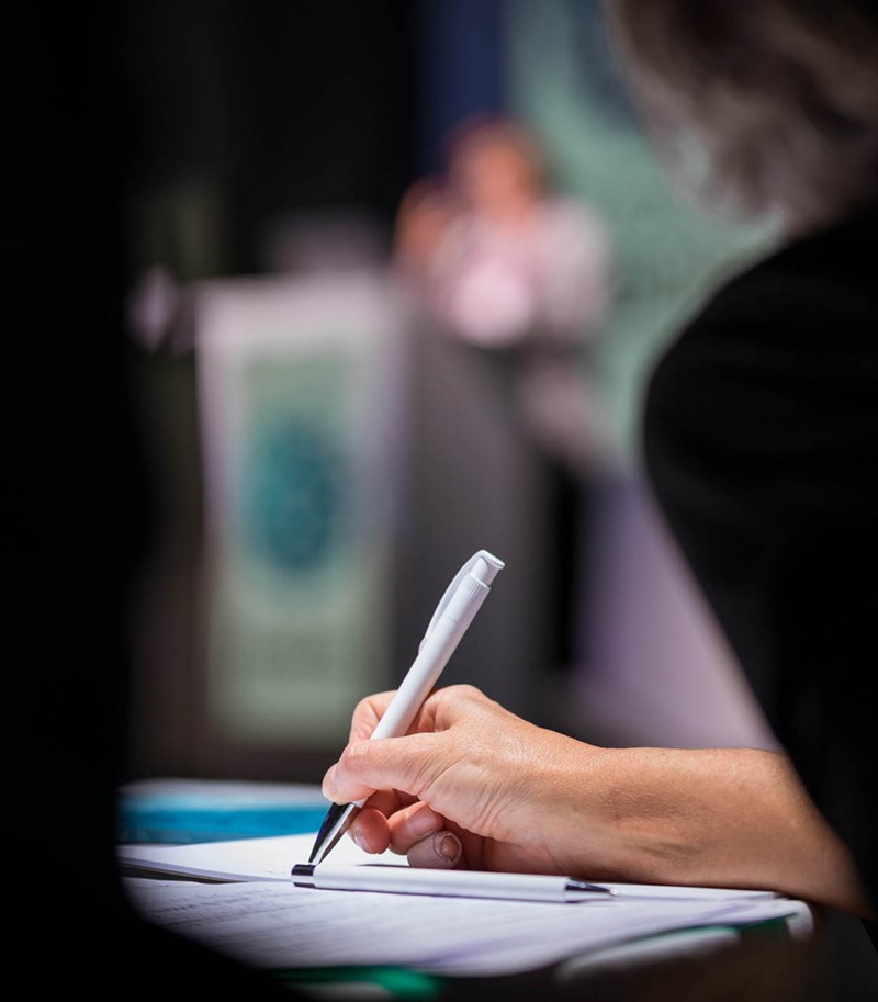 Woman's hand taking notes with white pen during a speech, only hand and pen in focus, rest of picture blurry