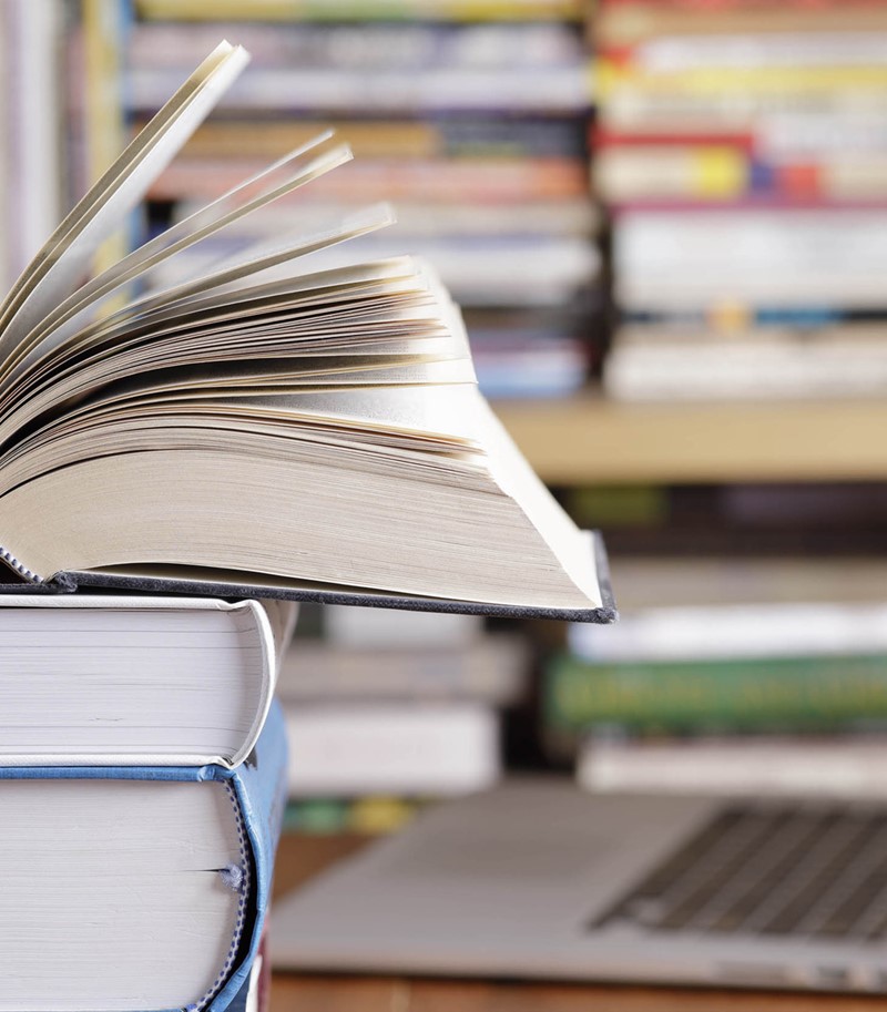 Three stacked books in focus in the foreground, blurred bookshelf and laptop on a desk in the background